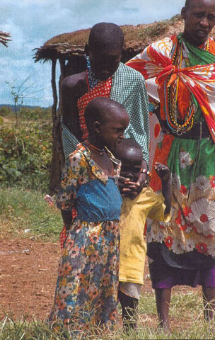 Two African adults and two young African children wearing colorful clothes are posing near a hut without looking at the camera. 