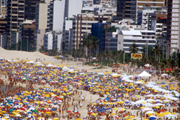 Aerial view of a beach crowded with tourists and mulitcoloured umbrellas with buildings in the background.