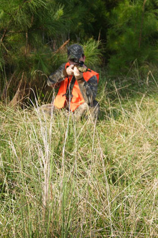 A man in a camouflage outfit with an orange vest is leaning in the grass, pointing his firearm towards the camera. 